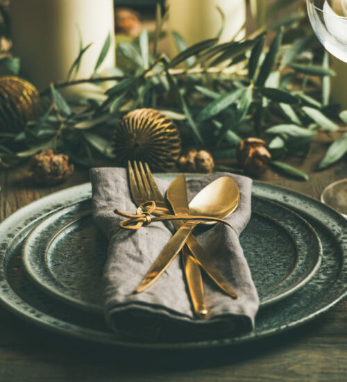 Christmas or New Years celebration party table setting. Plates, golden cutlery, glasses, gift box, festive branch decoration, candles, gliterring toys over rustic wooden table background, square crop